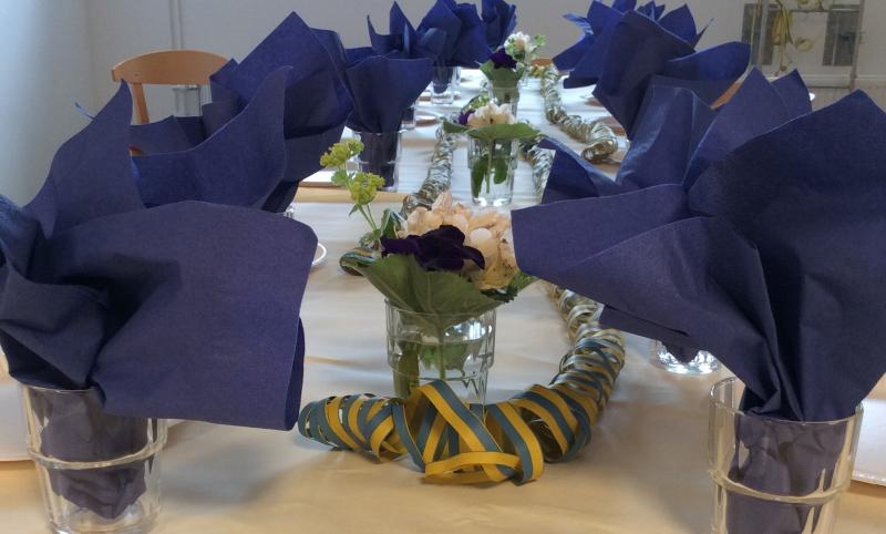 Bord dukat med blommor, blå-gula serpentiner och glas med blå servetter.
